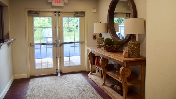 A hotel entrance with double doors, a wooden console table with lamps, a mirror, and a rug leading to the entrance. - Stay Plus Extended Stay Suites (Auburn)