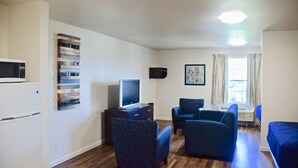 A compact living space with a kitchenette, a flat-screen TV, two blue armchairs, a small table, and a painting on the wall. - Stay Plus Extended Stay Suites (Auburn)