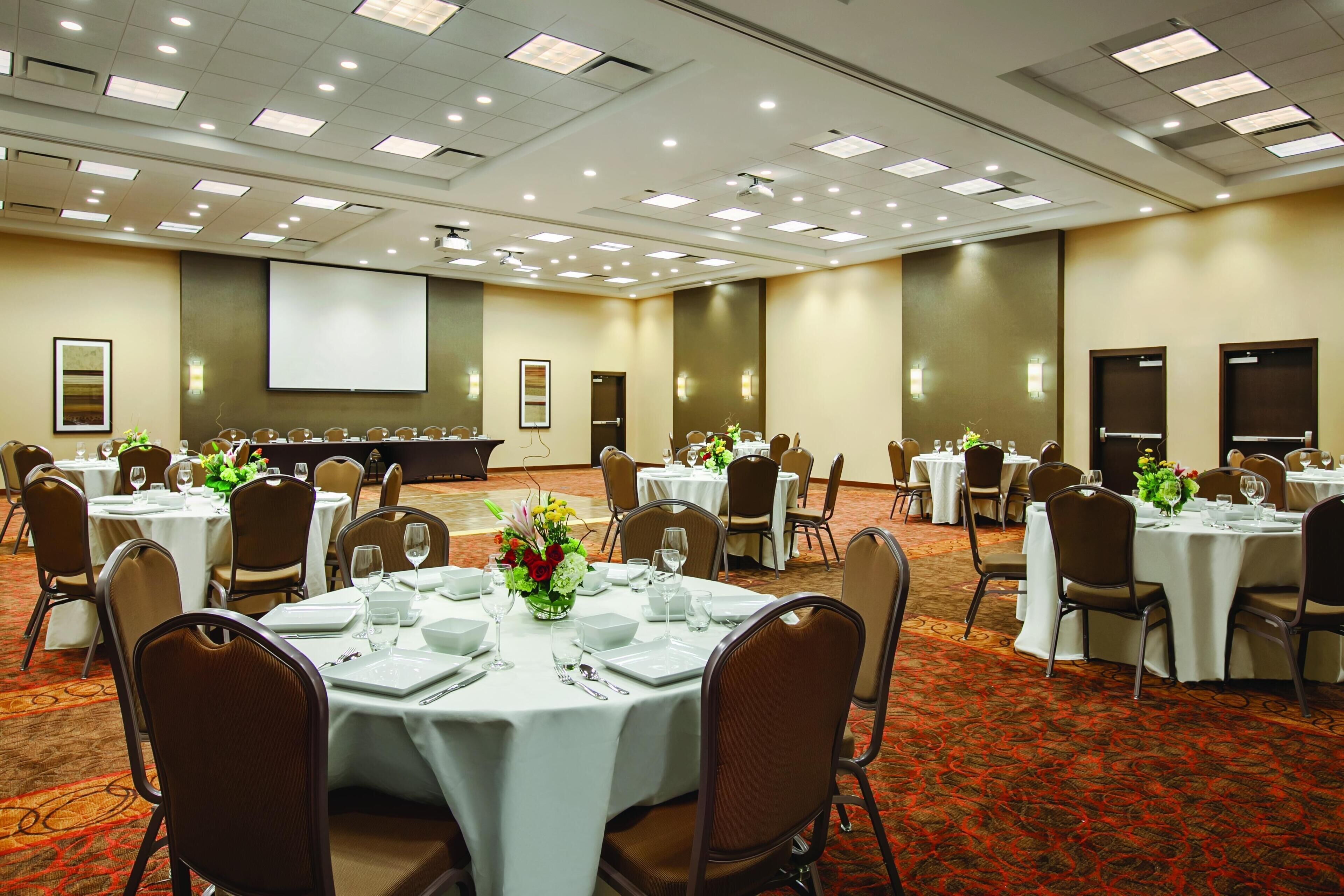 Un salón de conferencias con varias mesas redondas dispuestas para comidas, equipado con manteles blancos, sillas y centros de mesa florales.