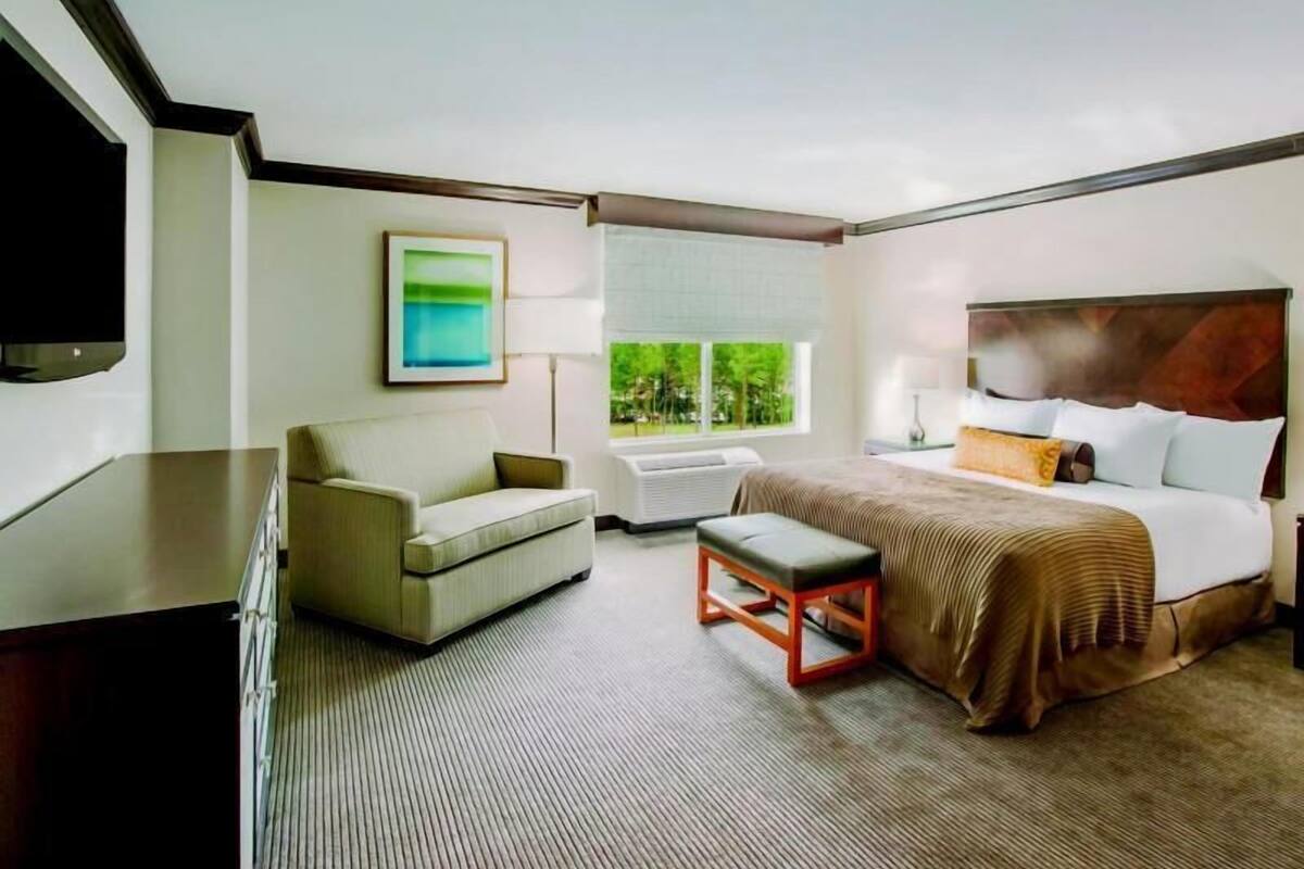 A hotel room with a bed, a sofa, a TV, a window with a view, and a painting on the wall.