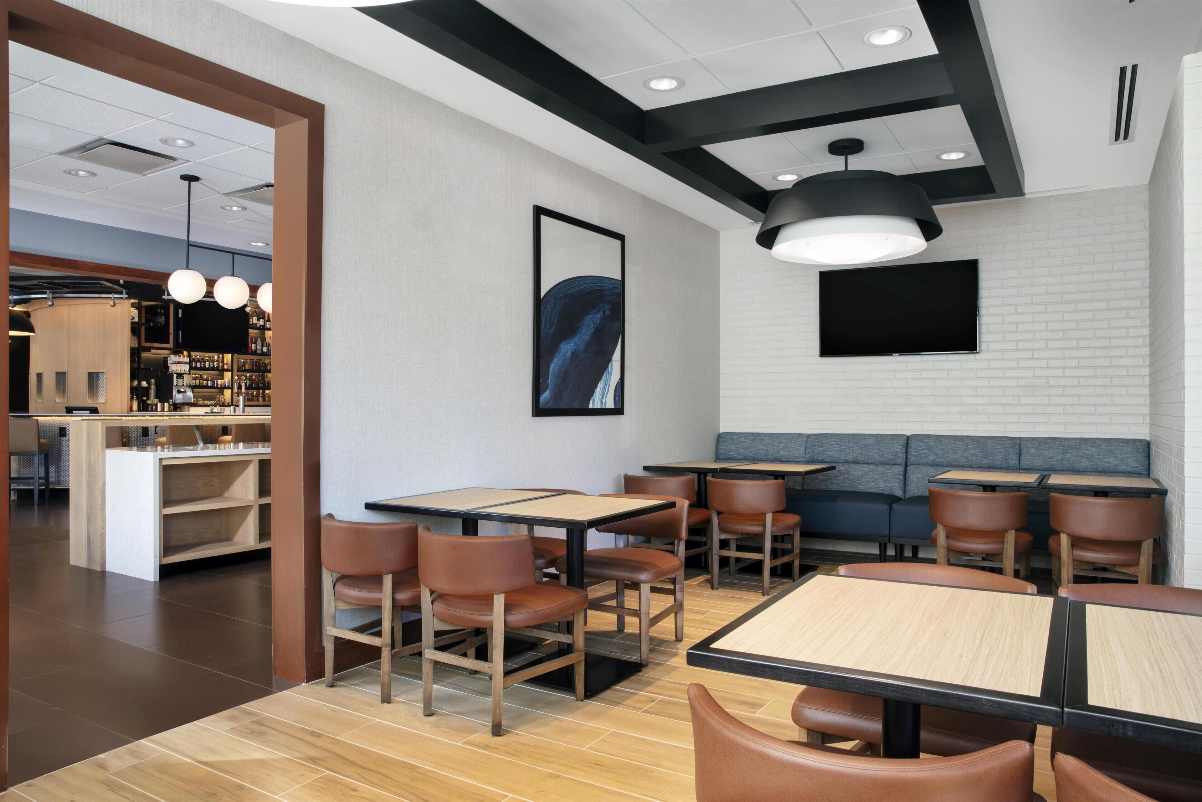 A modern restaurant interior with wooden tables and chairs, a bar area, and a large wall-mounted TV.