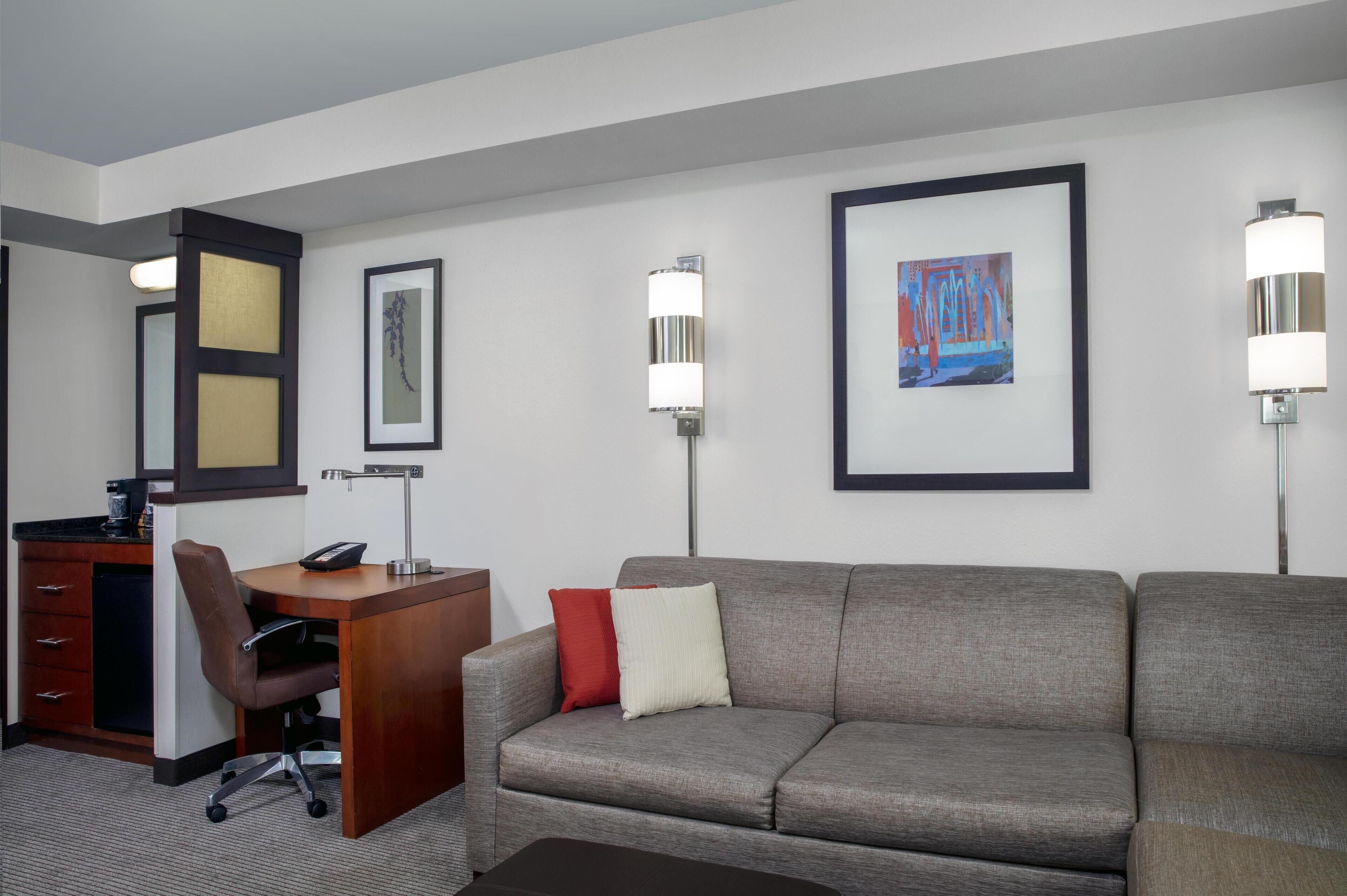 Habitación de hotel con escritorio, silla de oficina, sofá y dos lámparas de pared.