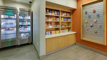 A kitchenette area with two stainless steel refrigerators stocked with beverages and snacks, a wooden cabinet with various food items, and a wall-mounted display with small packets.