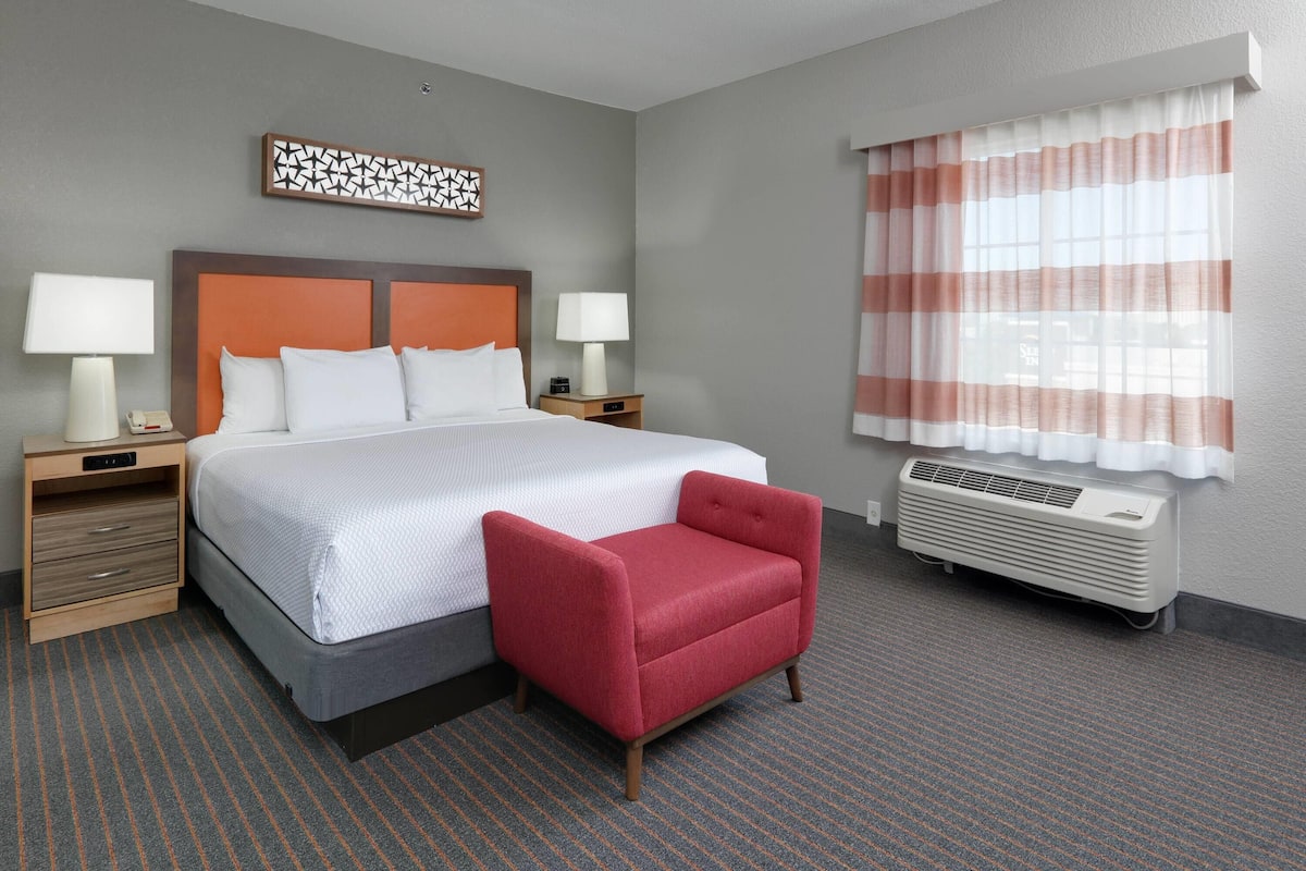 A hotel room with a large bed, two bedside tables with lamps, a red armchair, a decorative wall piece, and a window with striped curtains.