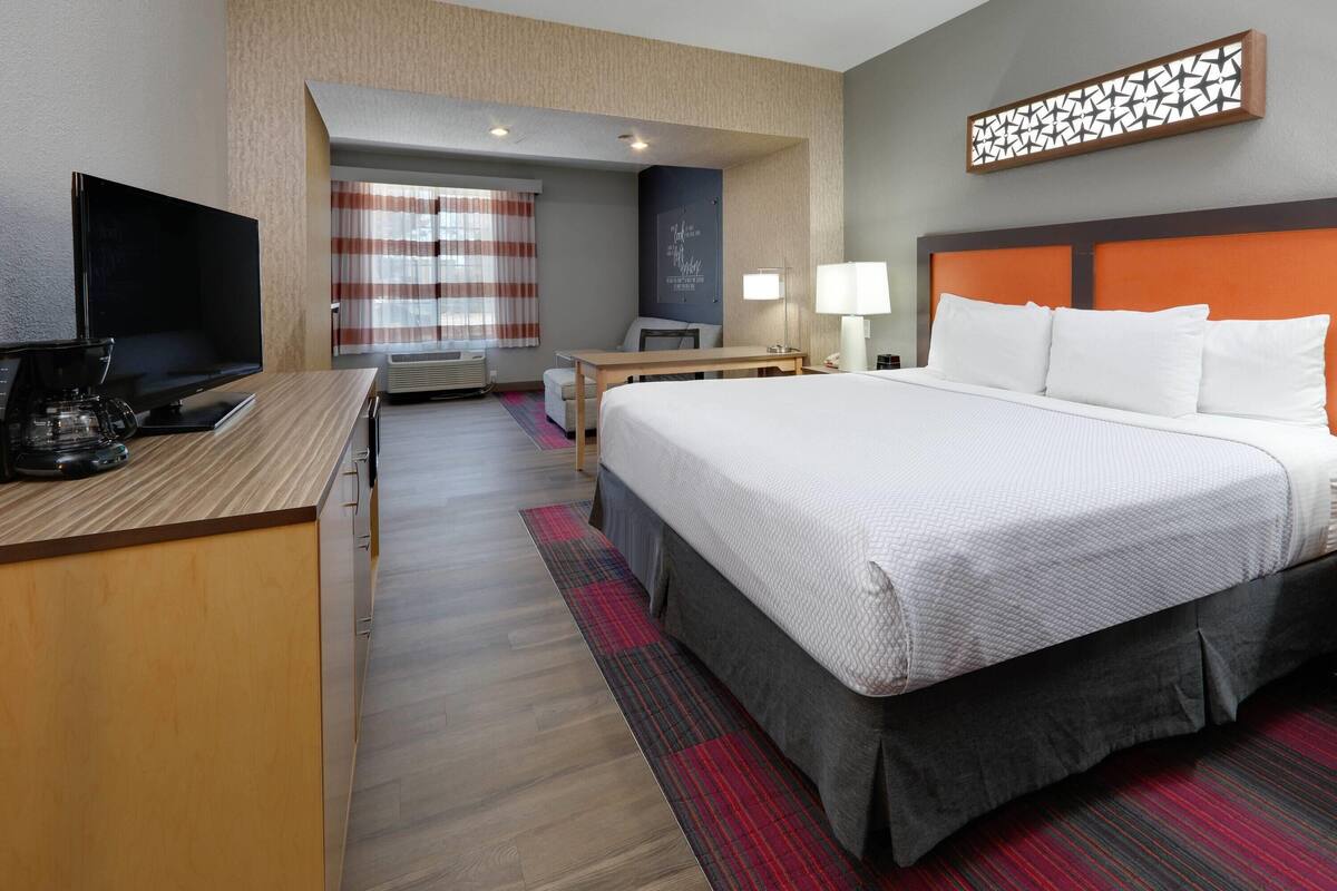 A hotel room with a bed, a desk with a coffee maker, a TV, a chair, and a window with curtains.