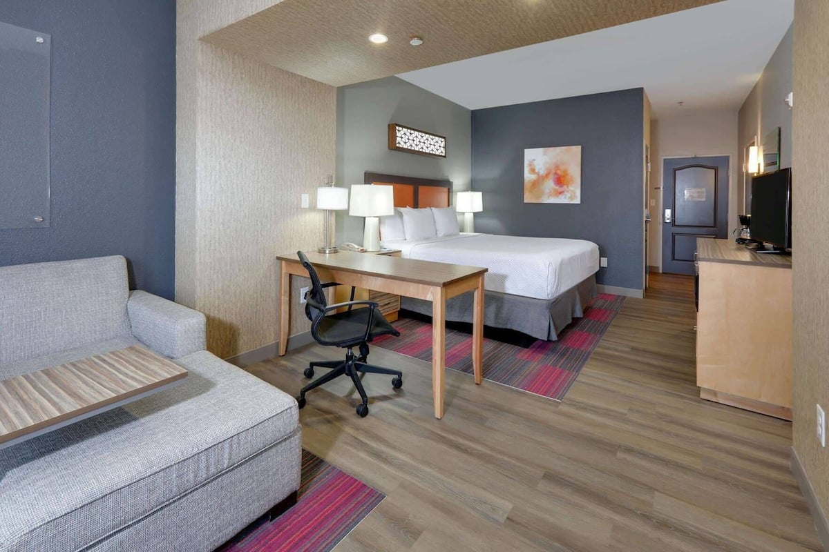 A hotel room with a bed, a desk with two lamps, a chair, a sofa, and a TV.