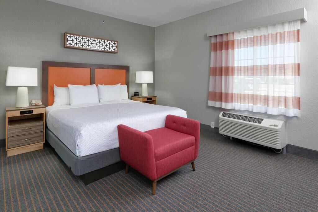 Una habitación de hotel con una cama grande, dos mesitas de noche con lámparas, un sillón rojo, un adorno en la pared y una ventana con cortinas a rayas.