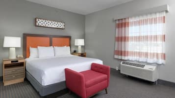 A hotel room with a large bed, two bedside tables with lamps, a red armchair, a decorative wall piece, and a window with striped curtains.