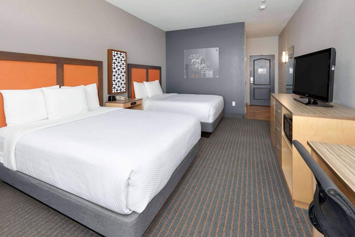A hotel room with two beds, a desk with a TV, a door, and a wall with a poster.