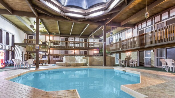 An indoor swimming pool with a tiled floor, wooden deck, and multiple seating areas with chairs and tables.