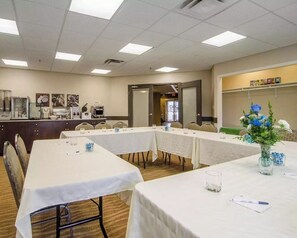 Un salón de conferencias con mesas largas, sillas y una zona de kitchenette.