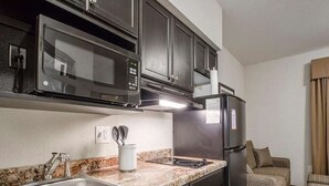 Cocina compacta con microondas, refrigerador, lavabo y un pequeño espacio de estar con sofá y reposapiés.