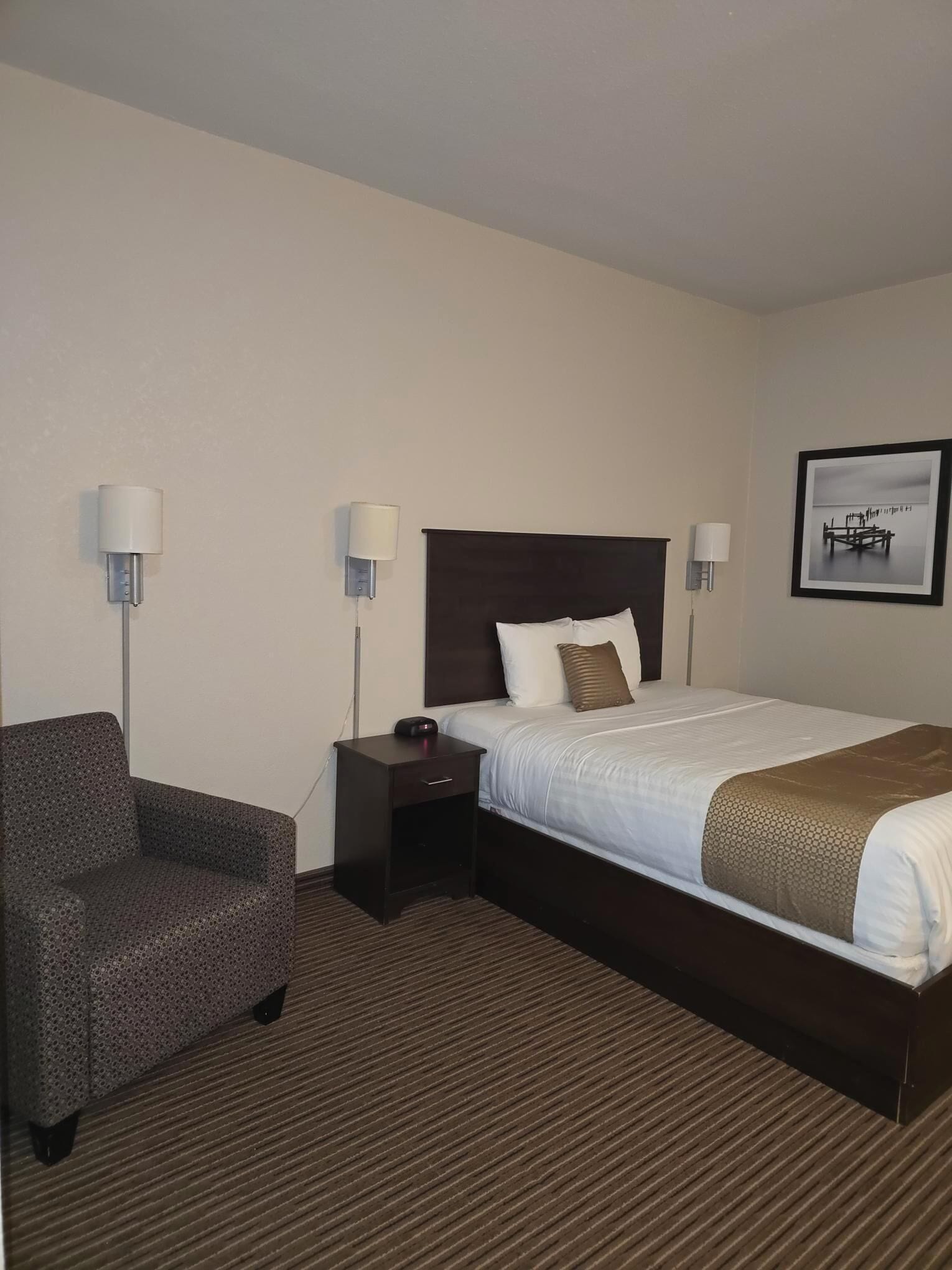 A hotel room with a bed, a chair, a nightstand, and two wall-mounted lamps.