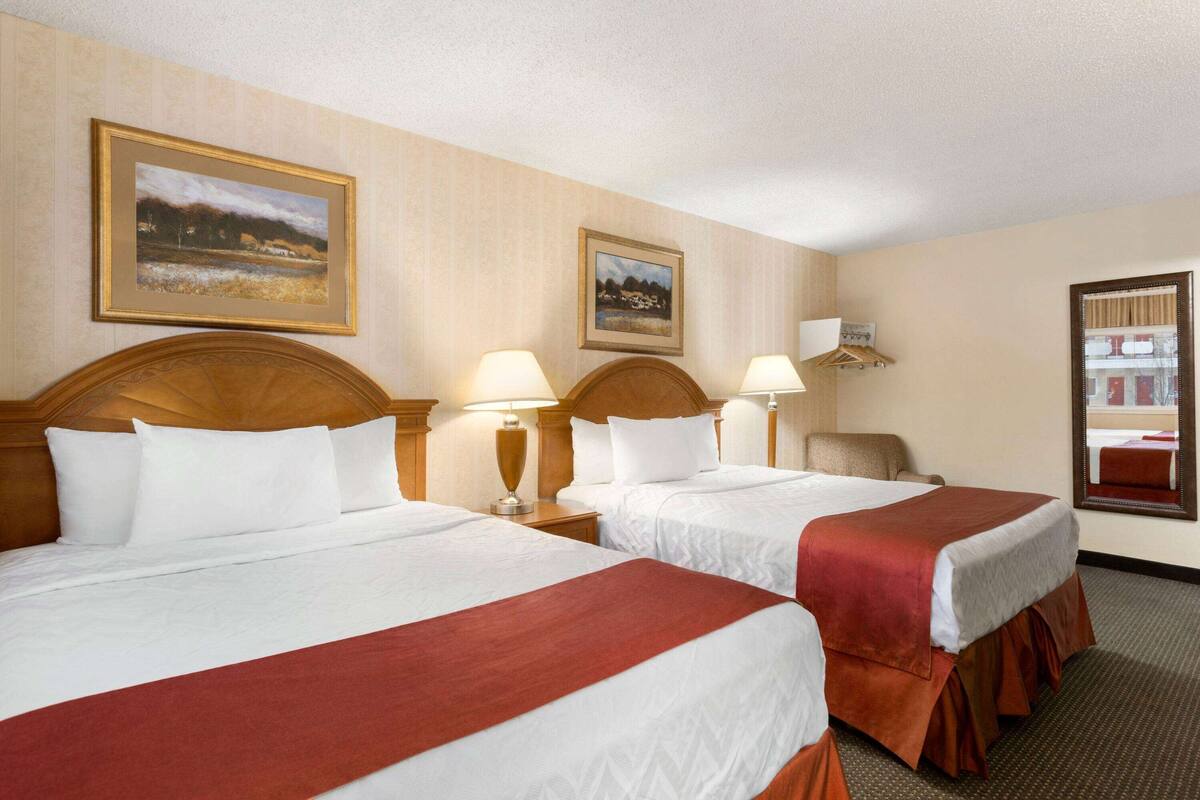 A hotel room with two beds, each with a wooden headboard, white bedding, and a red accent. There are two bedside lamps, a wooden nightstand, and a mirror on the wall.