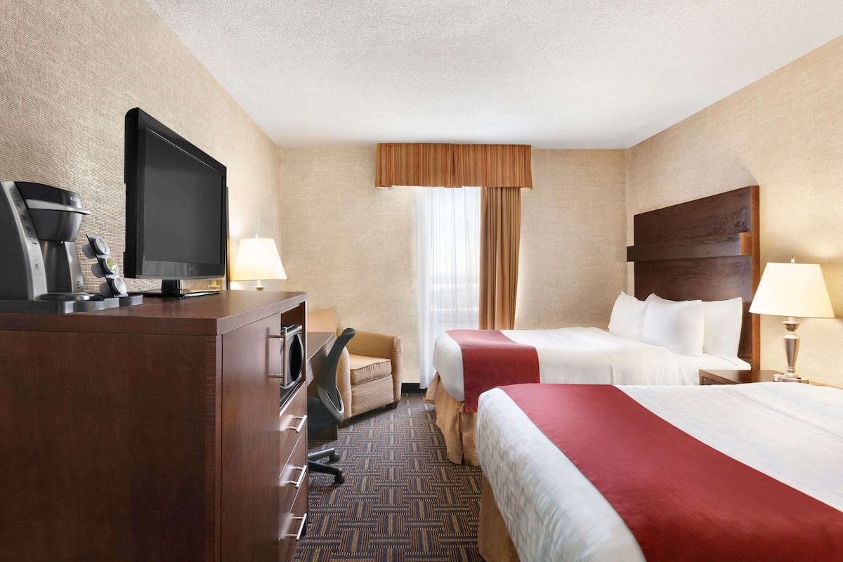 A hotel room with a TV, desk, chair, two beds, and a window with curtains.
