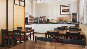 A hotel dining area with wooden tables and chairs, a kitchen in the background, and a framed picture on the wall. - Travelodge by Wyndham North Battleford (North Battleford)