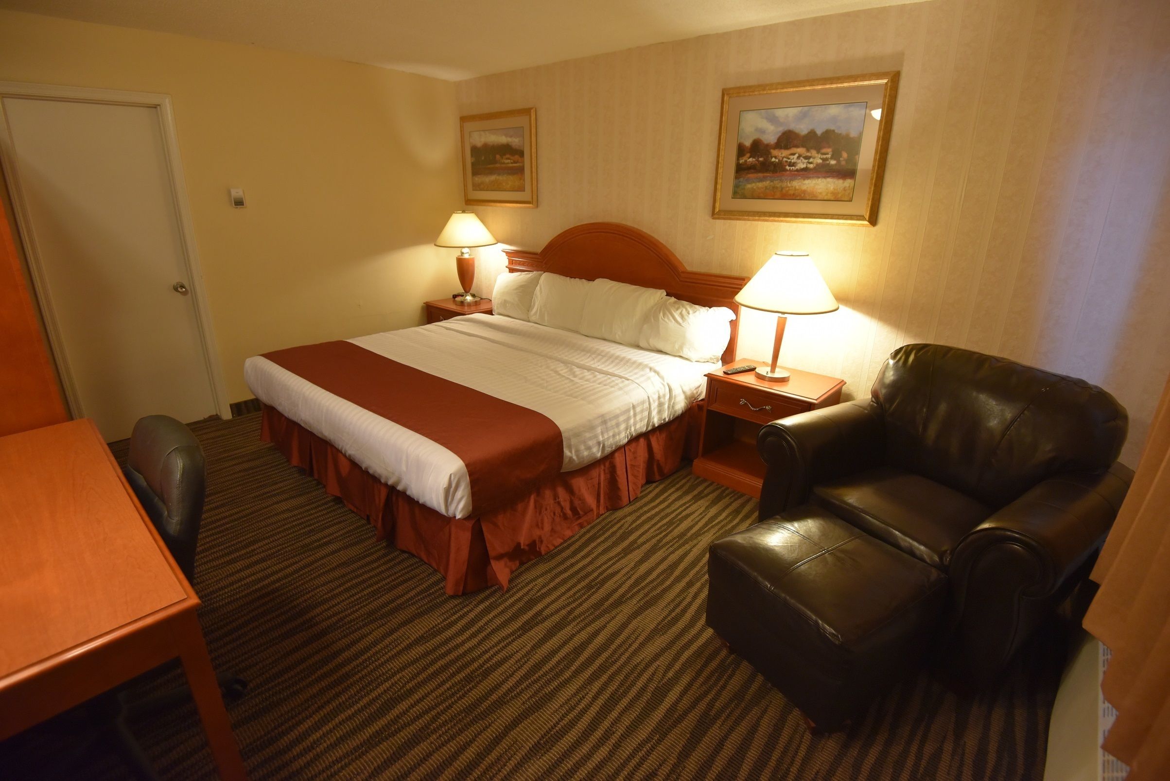 Quarto de hotel com uma cama, duas luminárias de cabeceira, uma poltrona de couro, uma escrivaninha de madeira e duas quadros na parede.