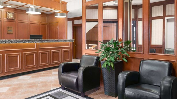 A reception area with a wooden reception desk, two black leather chairs, a potted plant, and a large mirror. - Travelodge by Wyndham North Battleford (North Battleford)