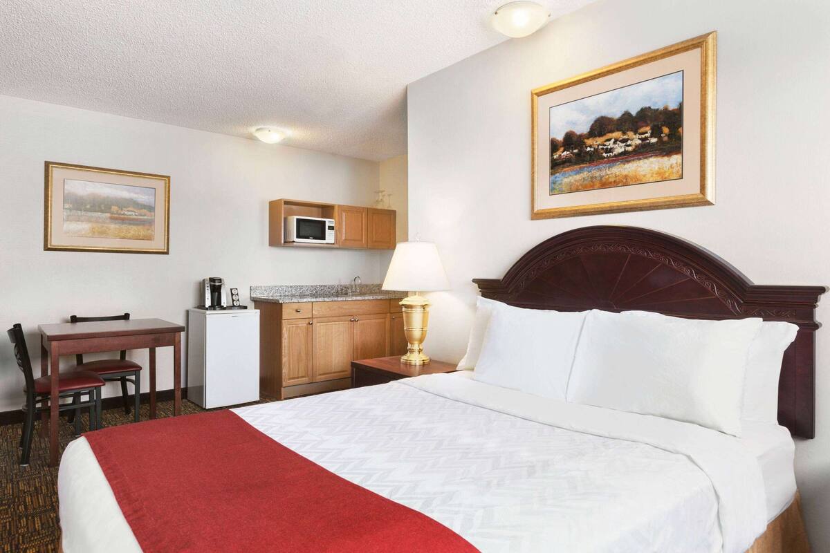 A hotel room with a bed, a desk, a chair, a microwave, and a lamp.