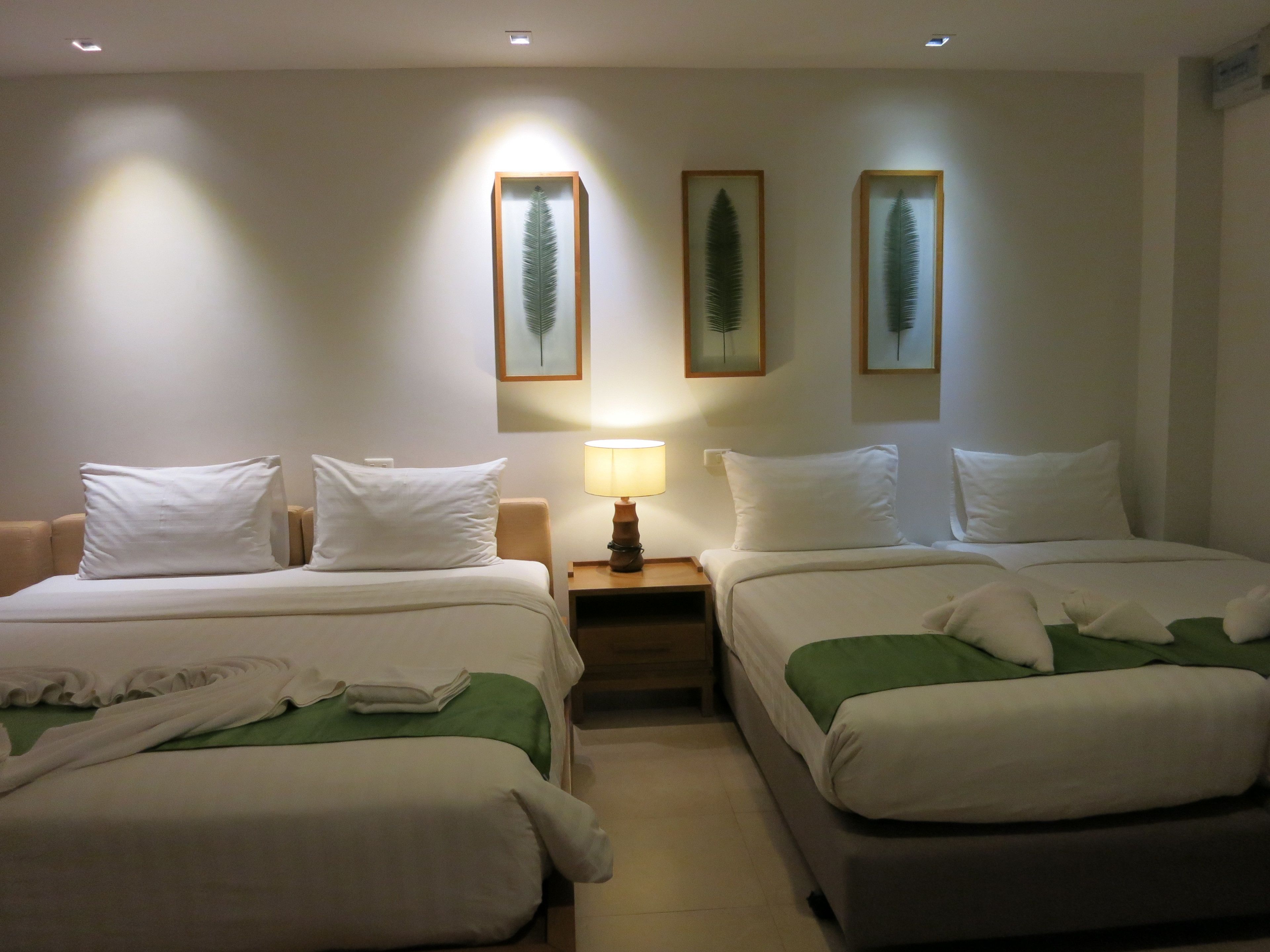 A hotel room with two single beds, a nightstand with a lamp, and three framed leaf wall art pieces.