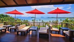 Eine Holzterrasse mit Außenbestuhlung und roten Sonnenschirmen mit Blick auf Strand und Meer.
