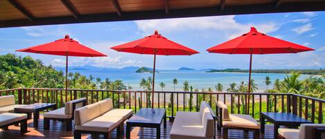 A wooden deck with outdoor seating and red umbrellas overlooking a beach and ocean.