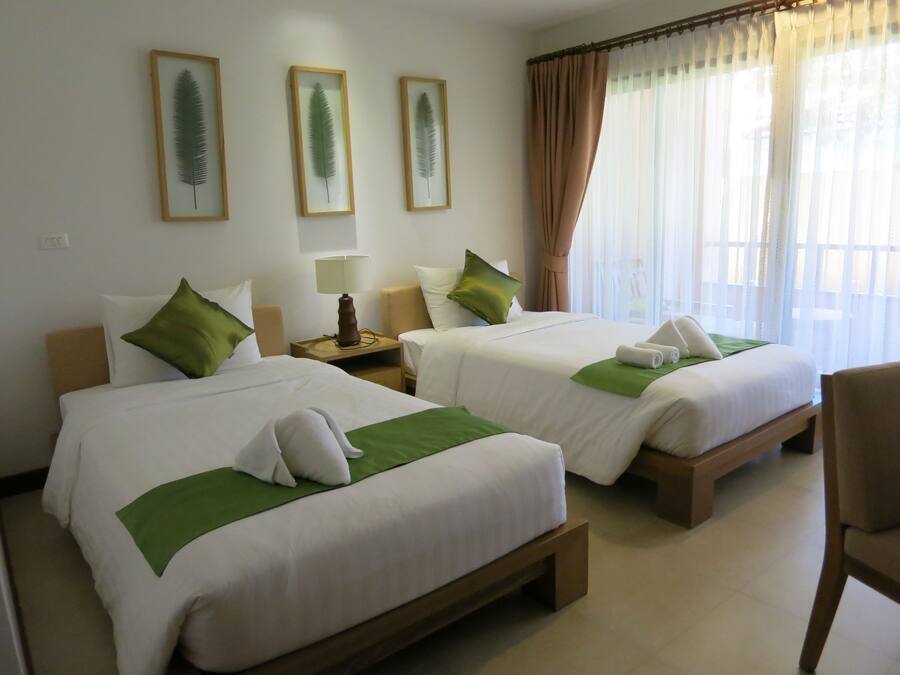 Quarto de hotel com duas camas, cada uma com almofadas verdes e um cobertor branco. Há folhas emolduradas na parede, um abajur em uma mesa de cabeceira de madeira e uma cadeira no canto.