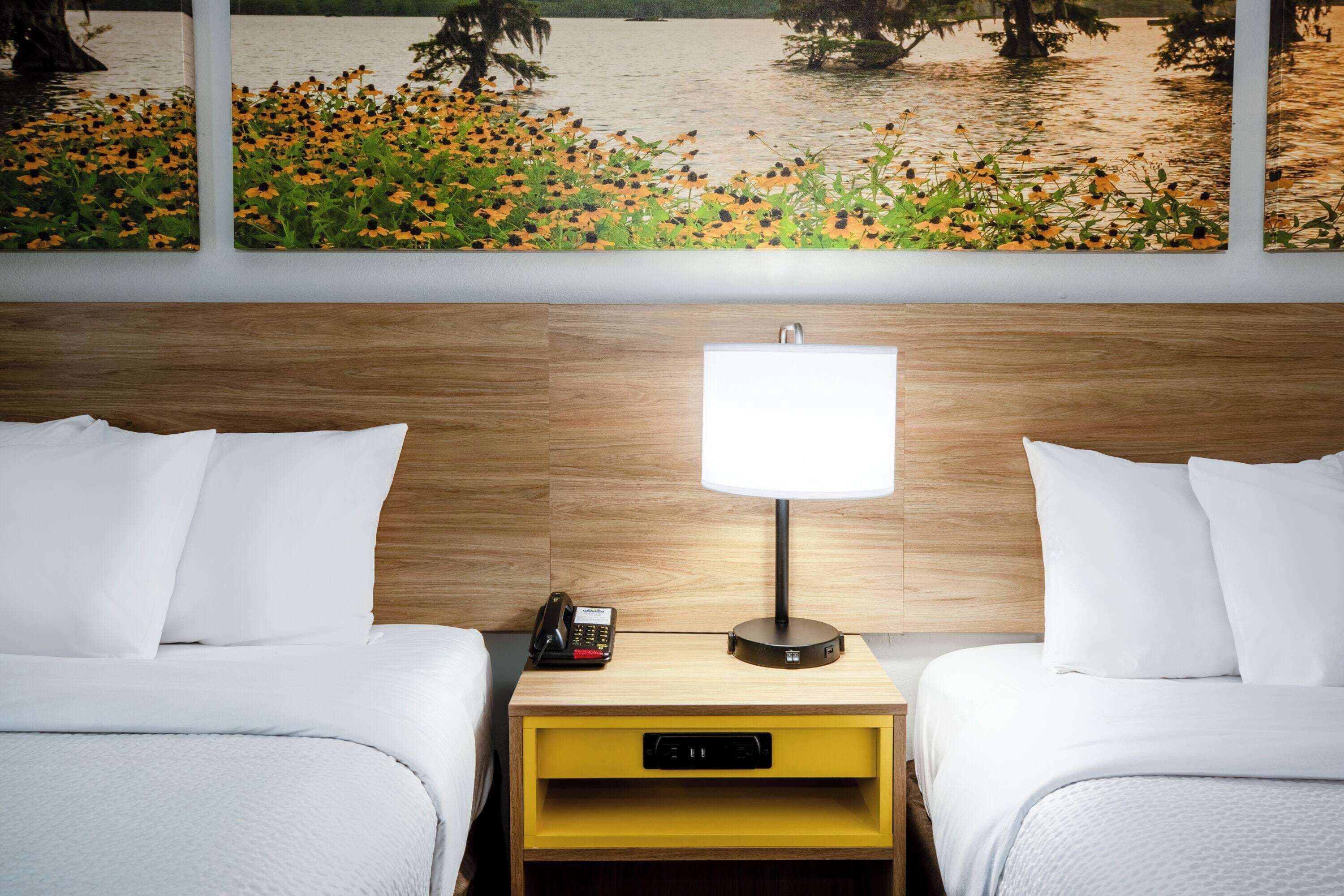 A hotel room with two beds, a nightstand with a lamp, a phone, and a small yellow drawer.