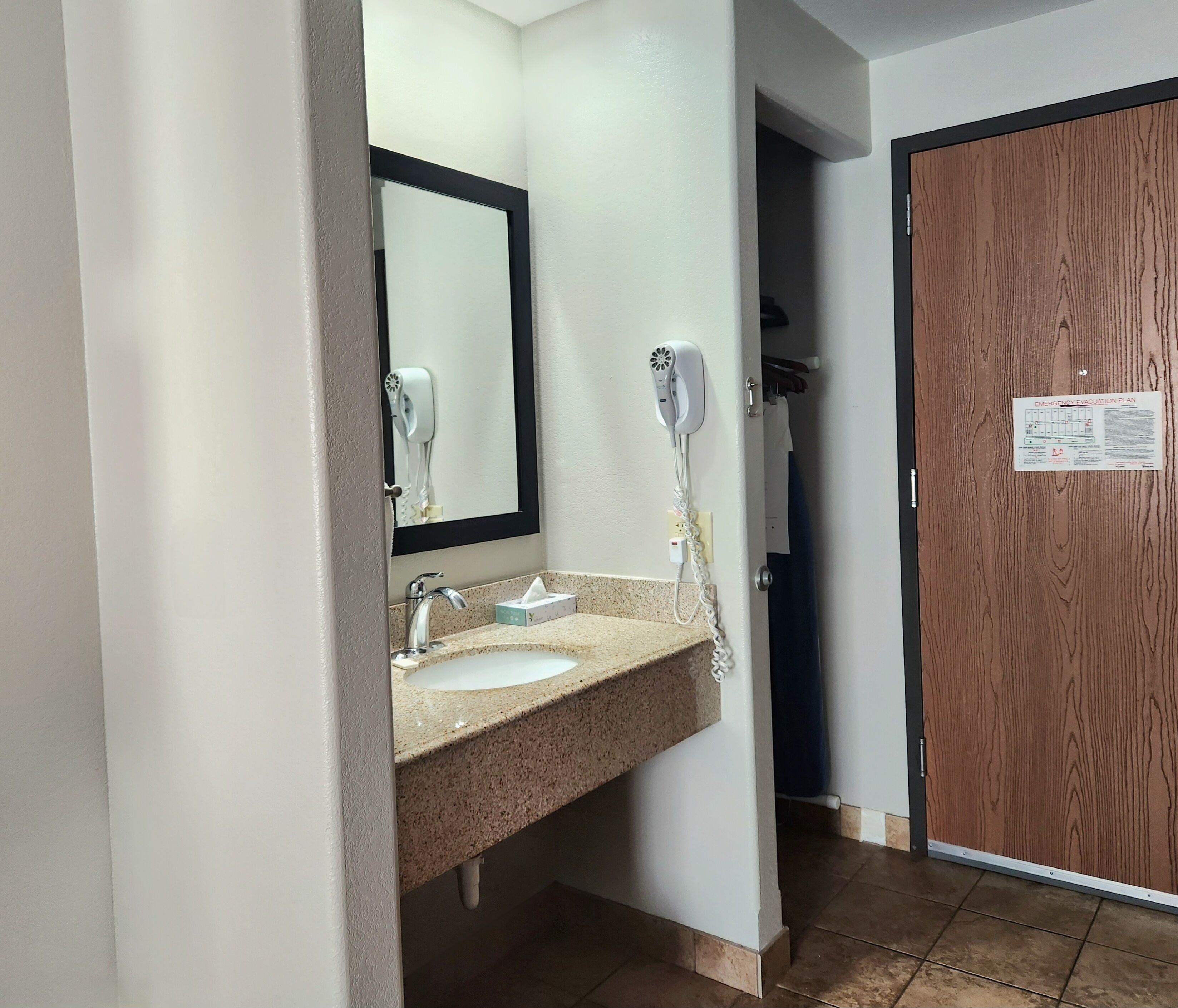 A bathroom with a sink, mirror, and hairdryer. A wooden door with a sign is visible in the background.