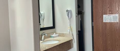 A bathroom with a sink, mirror, and hairdryer. A wooden door with a sign is visible in the background.