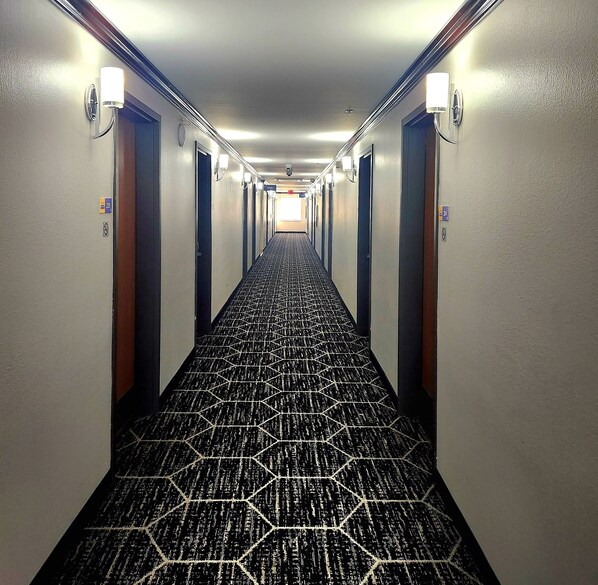 Pasillo de hotel con alfombra de estampado geométrico, puertas a ambos lados y lámparas fijadas a la pared.