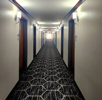 Pasillo de hotel con alfombra de estampado geométrico, puertas a ambos lados y lámparas fijadas a la pared.