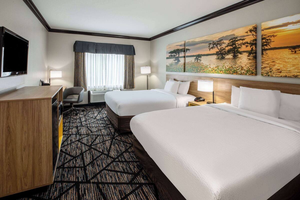 A hotel room with two beds, a desk, a chair, a TV, and a painting of a sunset over water.