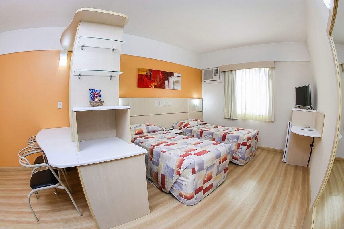 Quarto de hotel compacto com cama, escrivaninha e televisão.