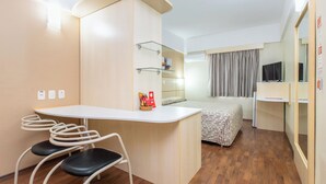 Een kleine hotelkamer met een kitchenette, een bed met een beddegoed met dessin, een bureau met stoelen, en een televisie.