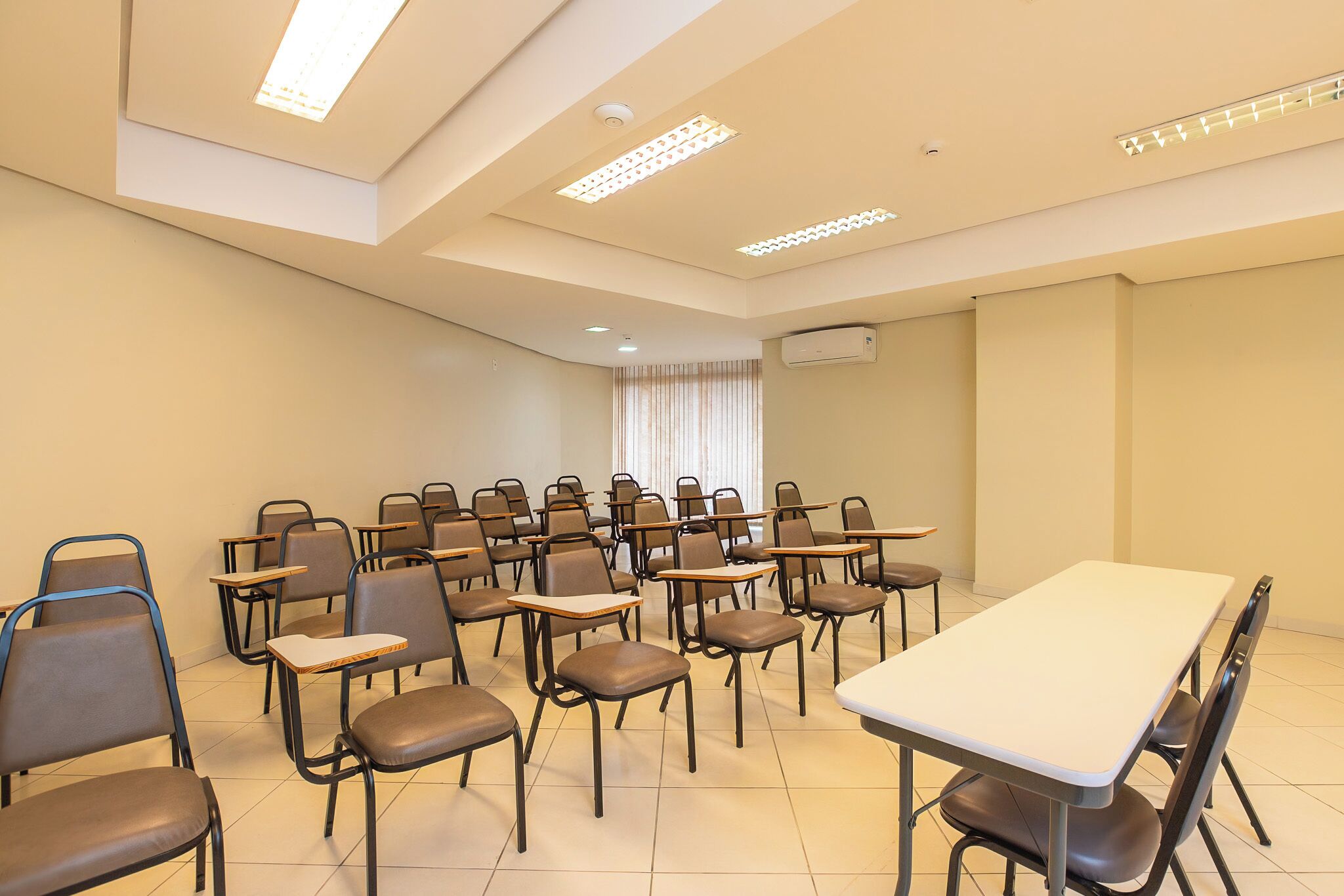 Sala de conferências com fileiras de cadeiras, uma mesa comprida e luminárias no teto.