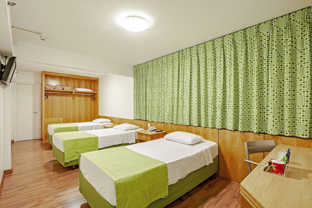 Ein Hotelzimmer mit zwei Einzelbetten, einem Holzschrank, einem Schreibtisch mit Stuhl und einem grün gemusterten Vorhang.