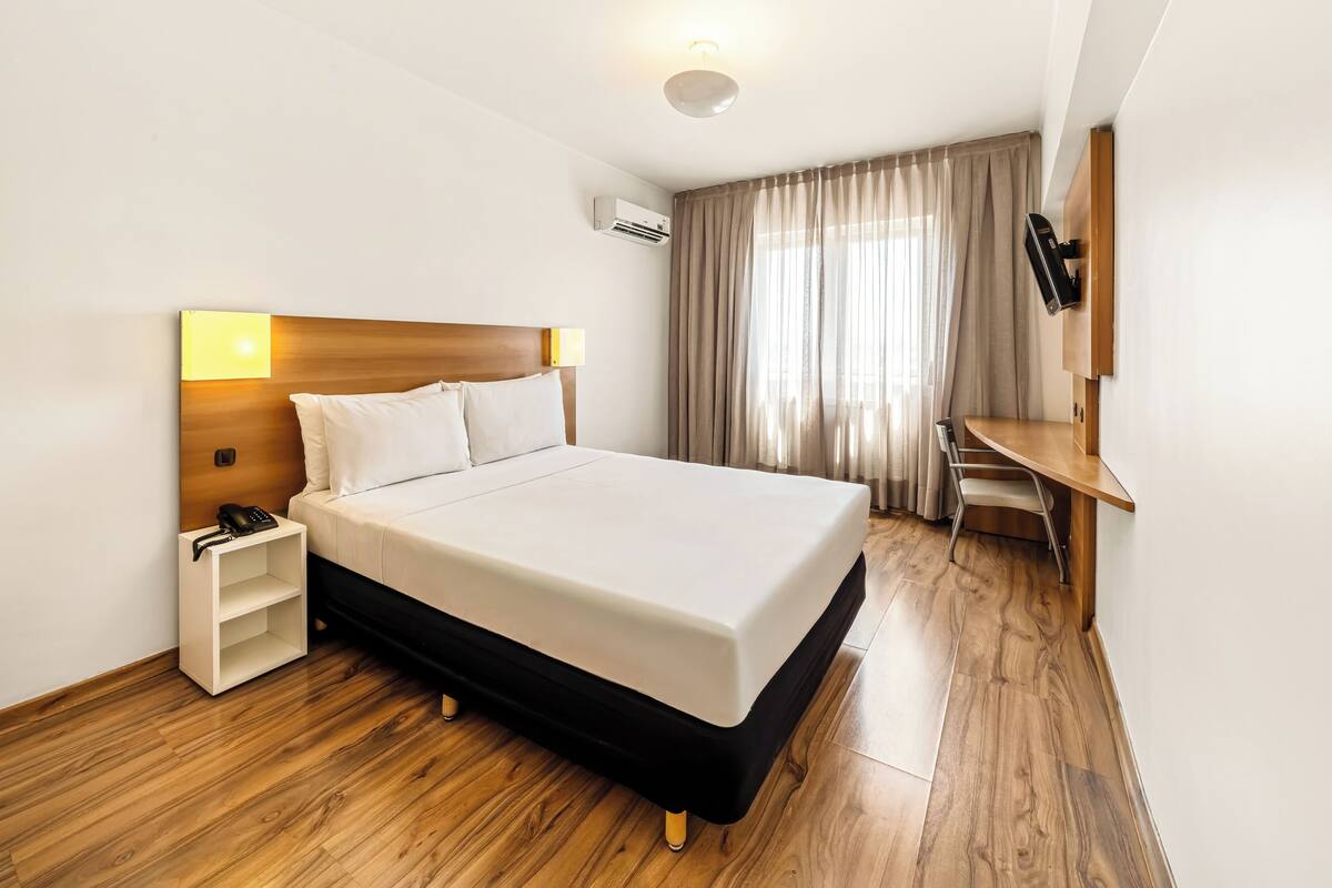 Ein modernes Hotelzimmer mit einem großen Bett, einem Nachttisch mit Lampe, einem Schreibtisch und einem an der Wand befestigten Fernseher.