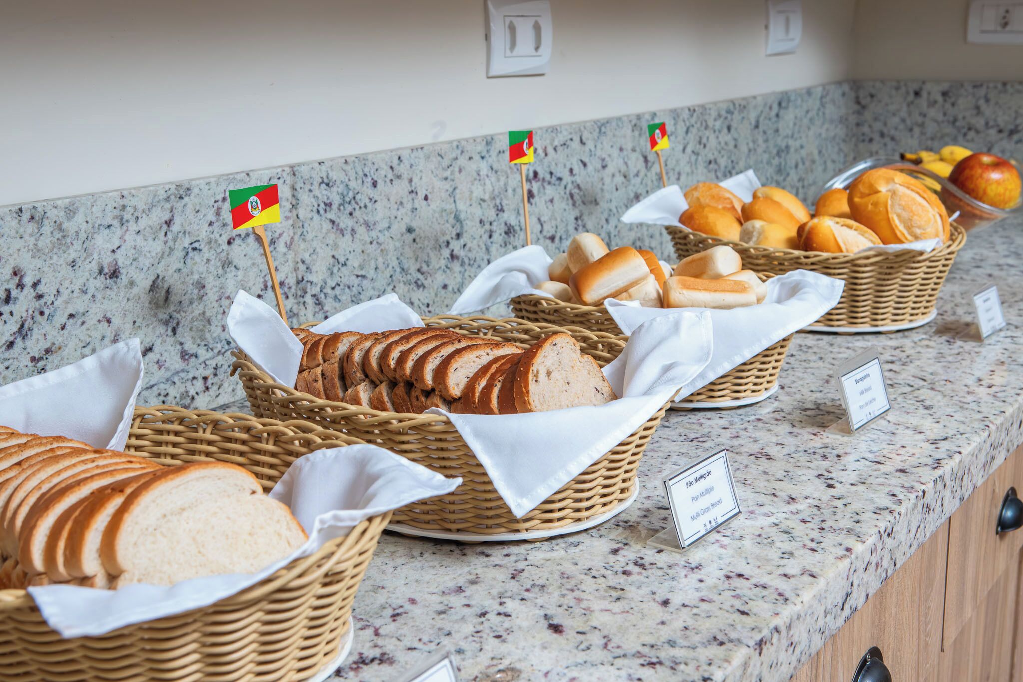 Buffet com diversos pães e doces, cada um identificado por uma pequena bandeira.