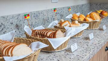 Buffet com diversos pães e doces, cada um identificado por uma pequena bandeira.