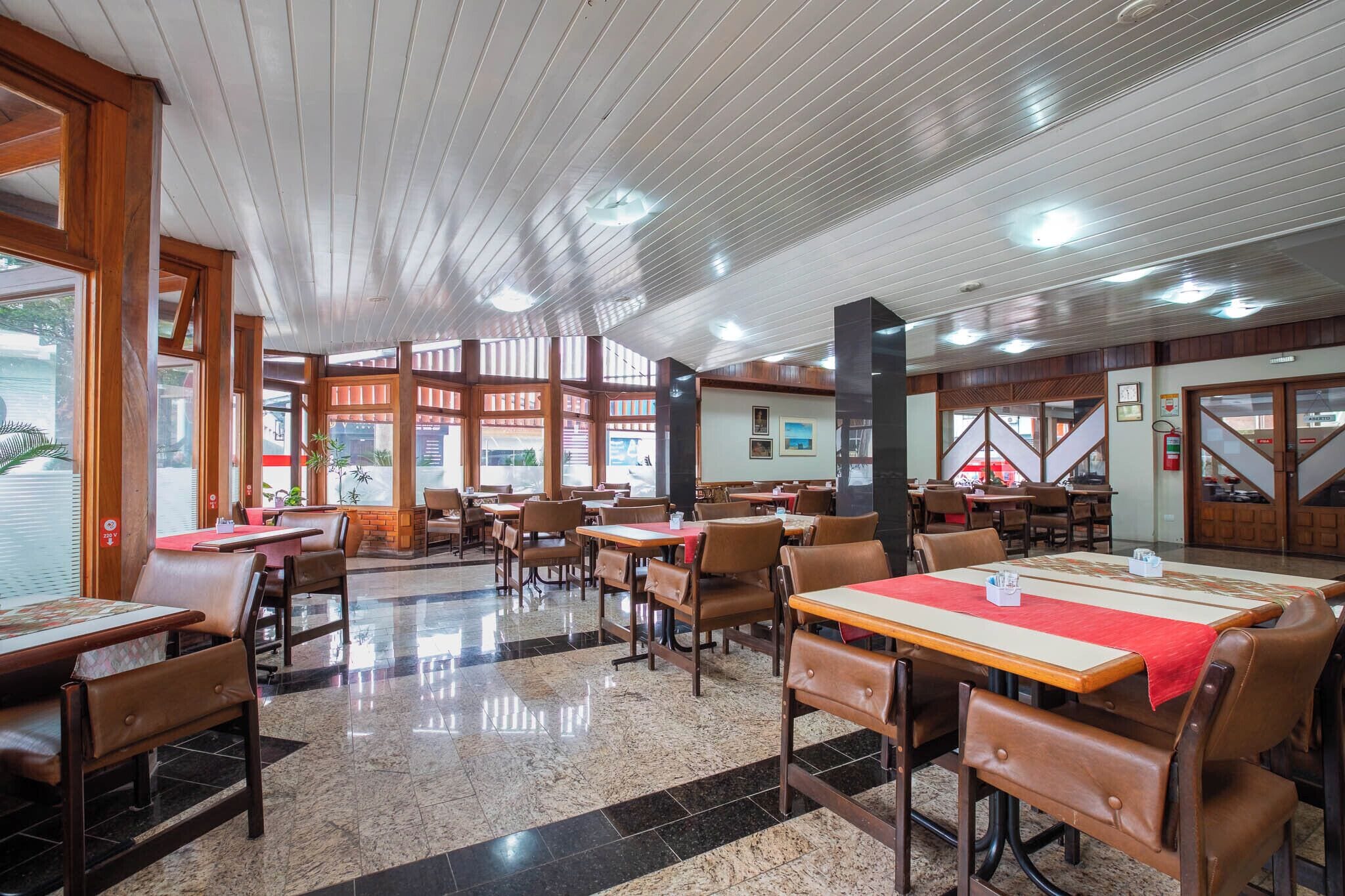 Um restaurante espaçoso com tetos altos, vigas de madeira e piso de mármore polido. As mesas estão dispostas para refeições, e há grandes janelas que permitem a entrada de luz natural.