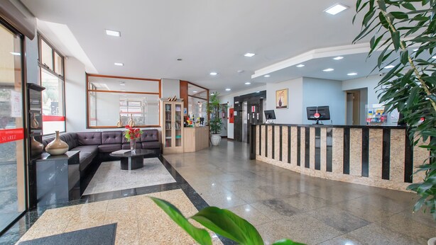 A hotel lobby with a reception desk, a sofa, a coffee table, and a potted plant. - Hotel Suárez São Leopoldo (Sao Leopoldo)