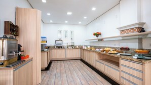 A modern kitchen with wooden cabinets, a stainless steel coffee machine, and a variety of food items on the counter. - Hotel Suárez São Leopoldo (Sao Leopoldo)