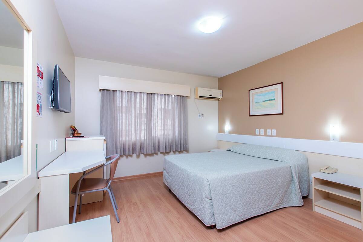Ein Schlafzimmer mit einem Bett, einem Schreibtisch, einem Stuhl, einer Klimaanlage, einem Fenster mit Vorhängen und einem Wandbild.