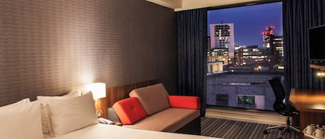 Een hotelkamer met een bed, een bank, een bureau, een stoel, een lamp en een groot raam met uitzicht op de stad.