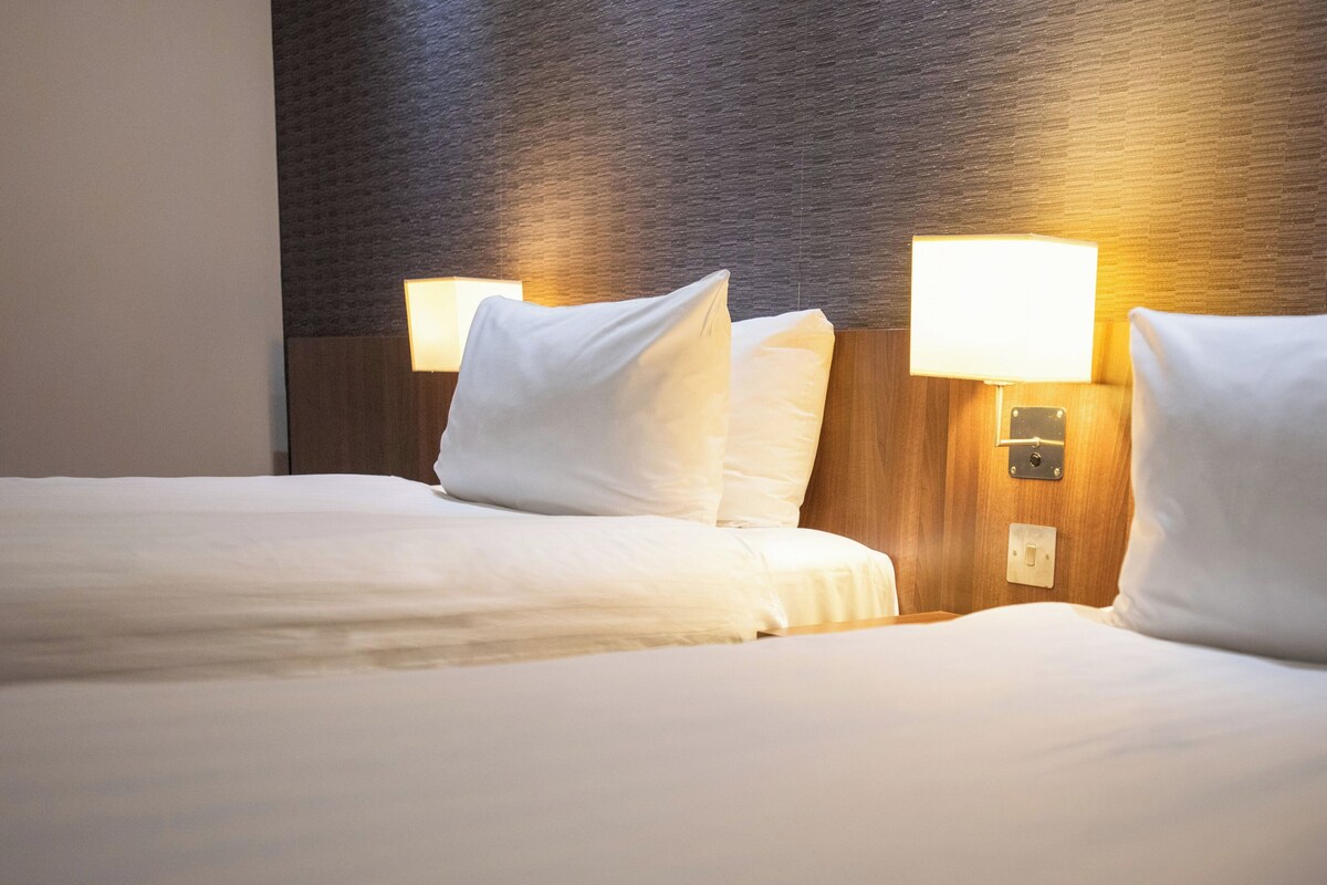 A hotel room with two beds, each with white linens and pillows, and two wall-mounted lamps.