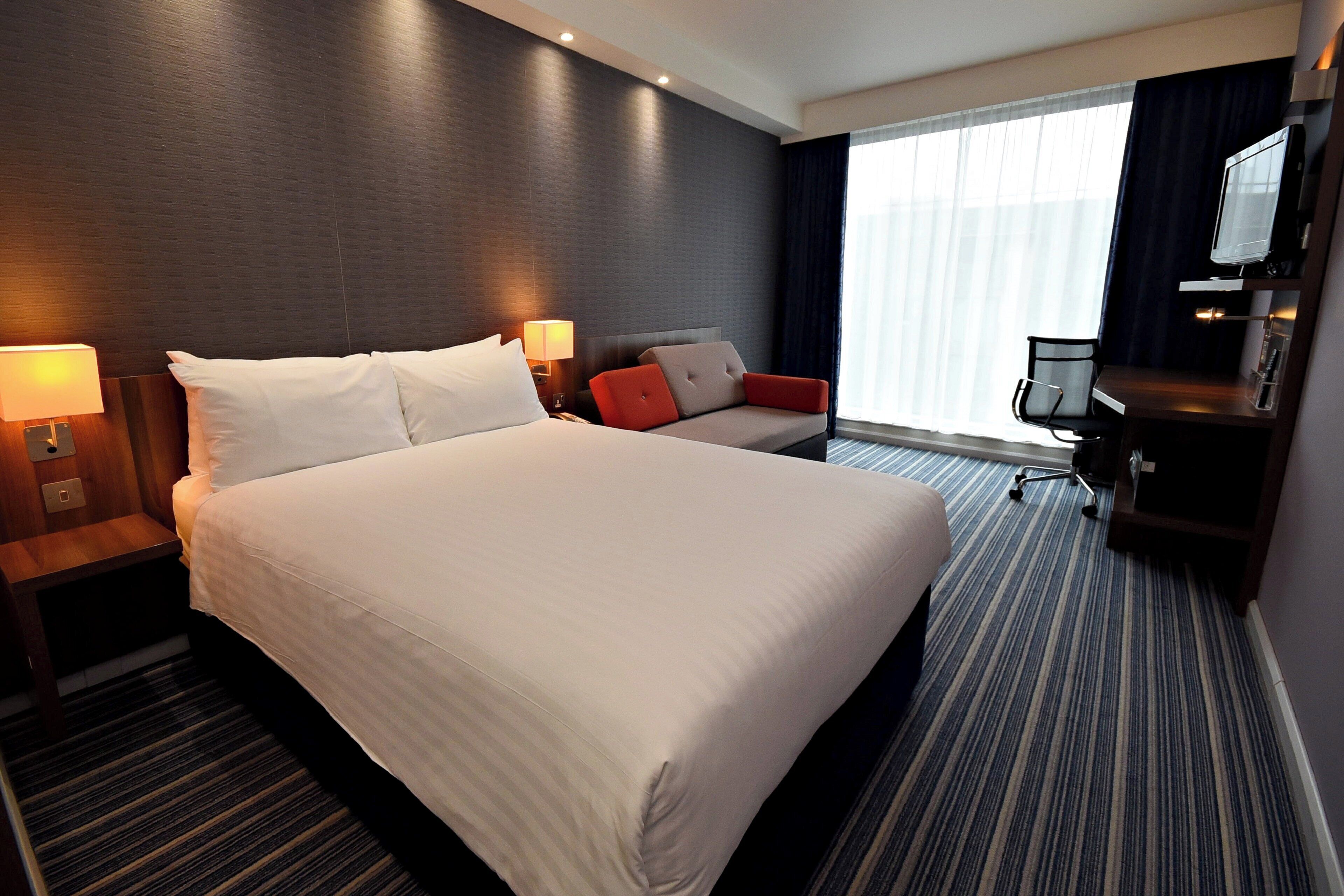 Habitación de hotel con una cama grande, un escritorio con silla, televisión y ventana con cortinas.