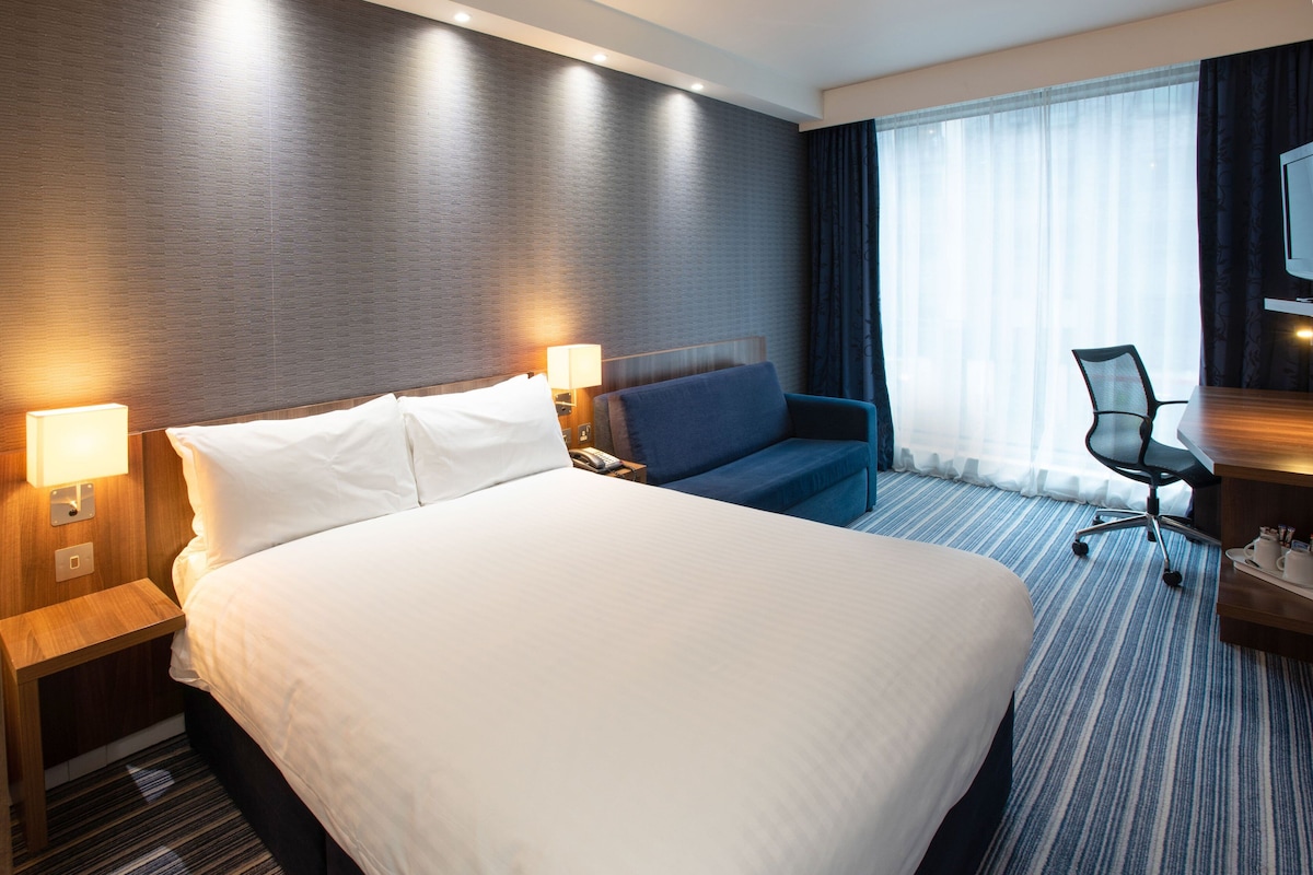 A hotel room with a large bed, a desk with a chair, a lamp, and a TV.
