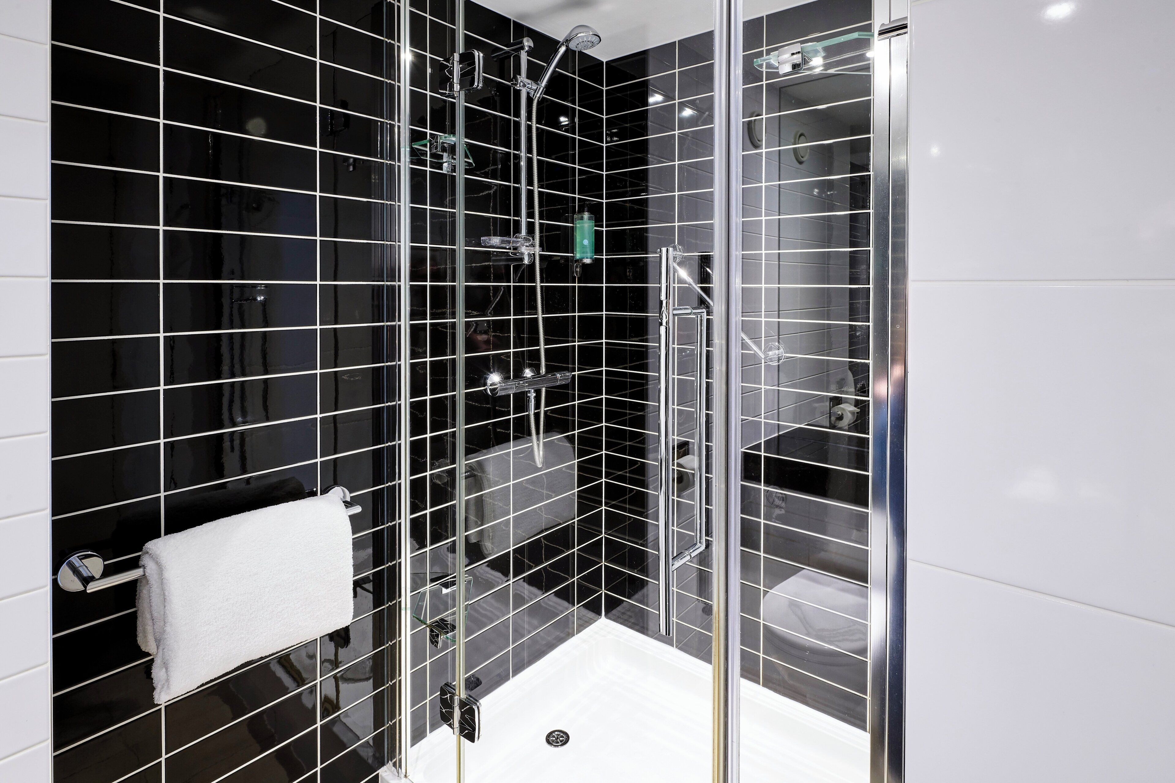 Une salle de bain moderne avec des murs recouverts de carreaux noirs, une douche en verre et une serviette blanche posée sur un porte-serviettes.