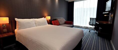 A hotel room with a large bed, a desk with a chair, a TV, and a window with curtains.